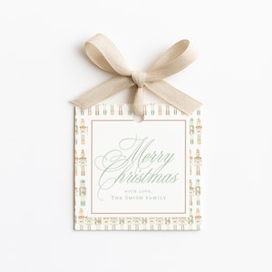 May include: A square Christmas gift tag with a beige ribbon bow. The tag has a border of toy soldier illustrations. The words "Merry Christmas" are written in elegant script, with "with love, The Smith Family" below.