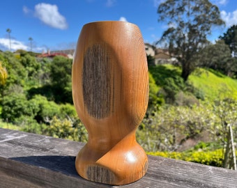 May include: A handcrafted wooden vase with a unique, curved design. The vase is made of light brown wood with darker, textured accents. The vase has a wide, rounded top and a flared base. The background features a sunny day with green hills and a blue sky.