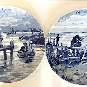 May include: Two blue and white Delftware plates depicting scenes from the Dutch countryside. The first plate shows a family in a boat on a canal, with the text "Juni" at the top. The second plate shows a hunter with a gun standing on a bridge, with the text "Oktober" at the top.