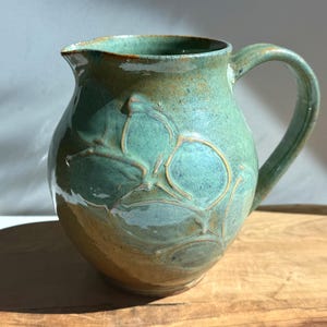 May include: A handmade ceramic pitcher with a rounded body and a curved handle. The pitcher is glazed in shades of teal and brown, with a leaf-like pattern on the front. The pitcher is sitting on a wooden surface.