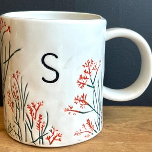 Pu&ograve; includere: Tazza in ceramica bianca con la lettera "S" nera e un design floreale rosso e verde. La tazza ha un manico grande ed &egrave; decorata con un motivo botanico. La tazza &egrave; su una superficie di legno.