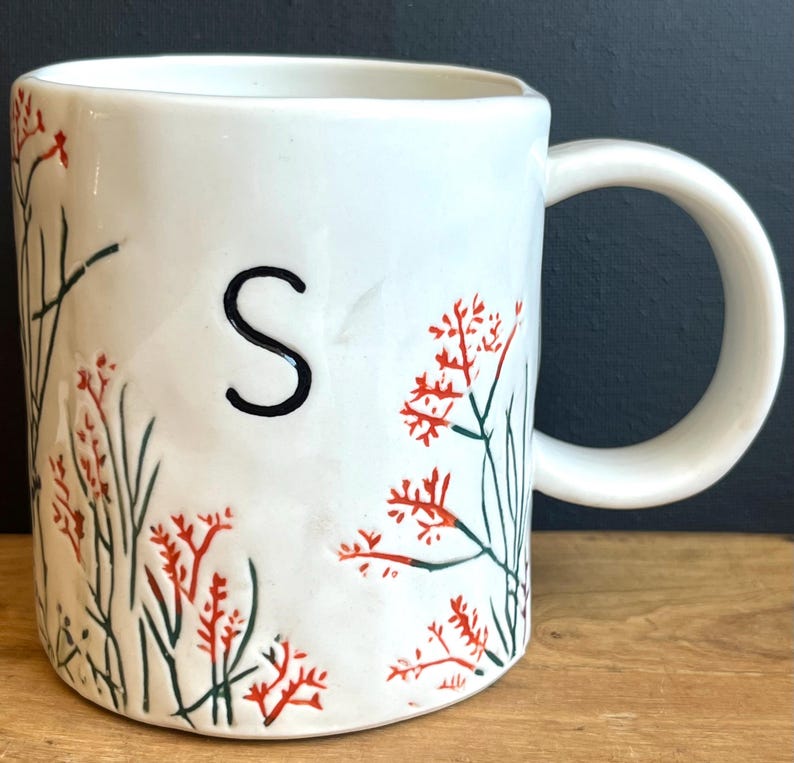 Pu&ograve; includere: Tazza in ceramica bianca con la lettera "S" nera e un design floreale rosso e verde. La tazza ha un manico grande ed &egrave; decorata con un motivo botanico. La tazza &egrave; su una superficie di legno.