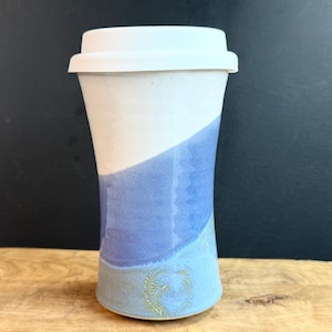 May include: A ceramic travel mug with a white lid. The mug is white on top, transitioning to a blue hue. A decorative emblem is near the base. The mug is on a wooden surface.