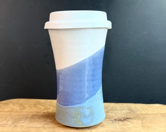 May include: A ceramic travel mug with a white lid. The mug is white on top, transitioning to a blue hue. A decorative emblem is near the base. The mug is on a wooden surface.