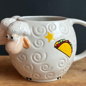 May include: White ceramic mug with a sheep design, featuring a 3D sheep head and embossed swirls. A yellow star and a taco graphic decorate the side. The mug has a large handle and is set on a wooden surface.
