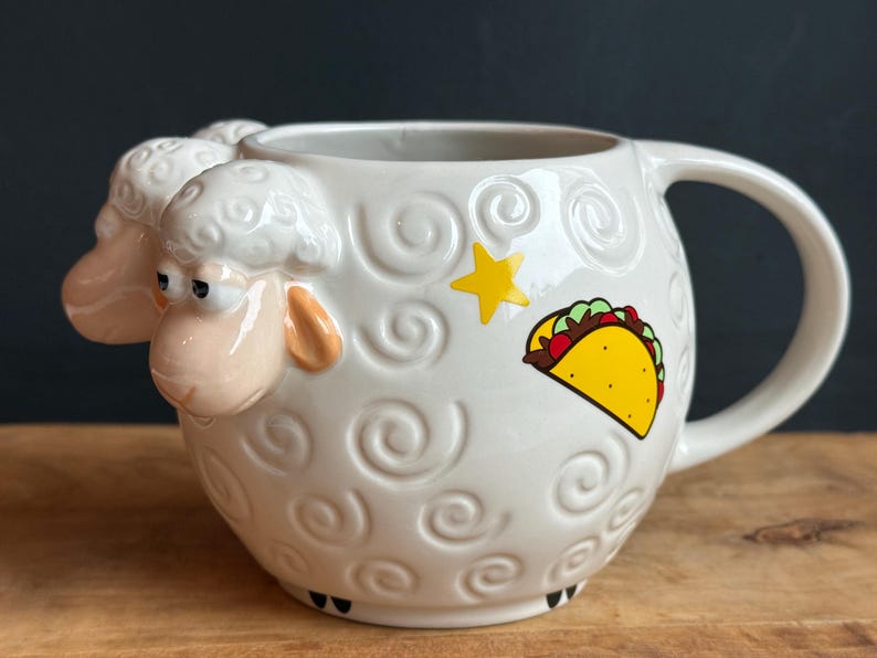 May include: White ceramic mug with a sheep design, featuring a 3D sheep head and embossed swirls. A yellow star and a taco graphic decorate the side. The mug has a large handle and is set on a wooden surface.