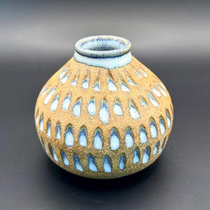 May include: A round, ceramic vase with a textured, tan exterior and a light blue interior and rim. The vase features a pattern of blue teardrop shapes across its surface. The vase is a decorative item.