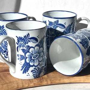 May include: Set of four white ceramic mugs with blue floral designs. The mugs have a classic shape and a comfortable handle.