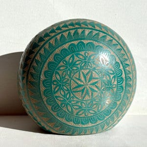 May include: A teal-colored, round decorative bowl with an intricate carved design. The bowl features a central starburst pattern surrounded by floral and geometric motifs. The carved details are highlighted with a contrasting tan color.