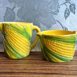 May include: A yellow ceramic pitcher and creamer set, each adorned with a detailed corn-on-the-cob design. The pitcher has a handle, and both pieces feature green husks. The set is displayed on a wooden tray.