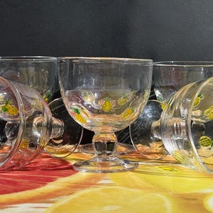May include: A collection of clear glass goblets with a decorative pineapple design. The goblets are arranged on a colorful surface with a black background. The pineapple design is yellow and green.