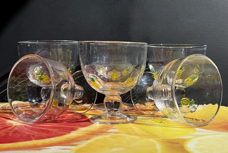 May include: A collection of clear glass goblets with a decorative pineapple design. The goblets are arranged on a colorful surface with a black background. The pineapple design is yellow and green.