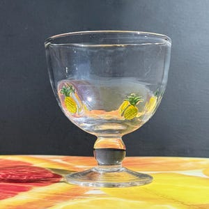 May include: A clear glass goblet with a short stem and a round bowl. The bowl is decorated with yellow and green pineapple designs. The stem has a black band. The goblet sits on a clear, round base.