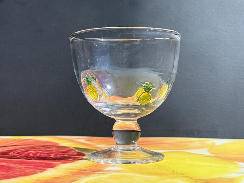 May include: A clear glass goblet with a short stem and a round bowl. The bowl is decorated with yellow and green pineapple designs. The stem has a black band. The goblet sits on a clear, round base.
