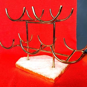 May include: A gold-coloured metal wine rack with a white and orange stone base. The rack is designed to hold multiple bottles, with curved holders. The background is a solid red.