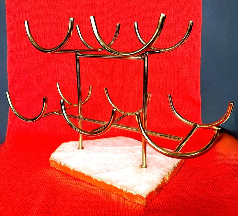 May include: A gold-coloured metal wine rack with a white and orange stone base. The rack is designed to hold multiple bottles, with curved holders. The background is a solid red.