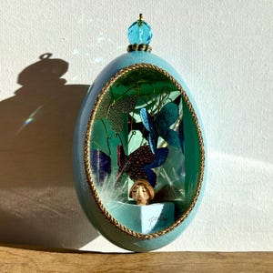 May include: A light blue, egg-shaped decorative piece with a gold rope trim. Inside, a miniature scene features butterflies, leaves, and a small figure. A blue crystal bead tops the piece. The word "poetry" is visible.