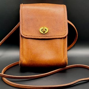 May include: A brown leather crossbody bag with a flap closure and a gold-tone clasp. The bag has a long, thin strap and a rectangular shape. The leather appears worn, giving it a vintage look. The bag is set against a dark background.