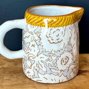 May include: A white ceramic pitcher with a rounded handle and a spout. The pitcher is decorated with a floral pattern in light blue and brown. The rim is accented with a yellow and brown striped design. The pitcher is on a wooden surface.