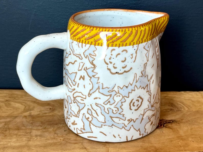 May include: A white ceramic pitcher with a rounded handle and a spout. The pitcher is decorated with a floral pattern in light blue and brown. The rim is accented with a yellow and brown striped design. The pitcher is on a wooden surface.