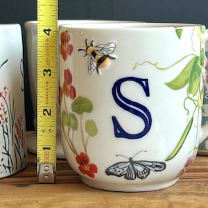 Pu&ograve; includere: Tazza in ceramica bianca con la lettera "S" blu, un'ape e una farfalla. La tazza &egrave; alta circa 7,6 cm. Disegni floreali e botanici decorano la tazza, insieme a un manico. Altre tazze con disegni floreali sono sullo sfondo.