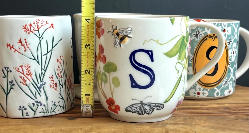 Pu&ograve; includere: Tazza in ceramica bianca con la lettera "S" blu, un'ape e una farfalla. La tazza &egrave; alta circa 7,6 cm. Disegni floreali e botanici decorano la tazza, insieme a un manico. Altre tazze con disegni floreali sono sullo sfondo.