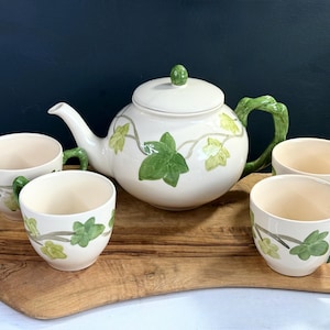 May include: A white ceramic tea set with green ivy leaf designs. The set includes a teapot with a green handle and lid, and four matching teacups with green handles. The set is displayed on a wooden serving tray.