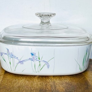 May include: White oval casserole dish with a clear glass lid. The dish features a floral design of purple irises and green leaves. The dish has two handles and is suitable for baking.