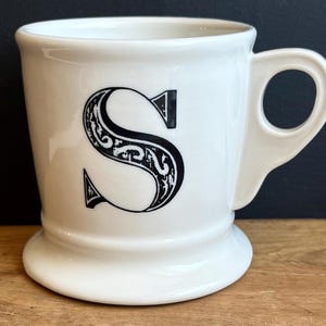 Pu&ograve; includere: Tazza in ceramica bianca con una grande lettera "S" nera e ornata sul davanti. La tazza ha una base arrotondata, un manico curvo e una finitura lucida. &Egrave; appoggiata su una superficie di legno.