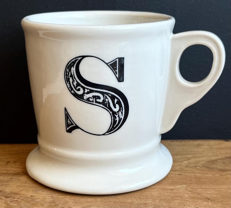 Pu&ograve; includere: Tazza in ceramica bianca con una grande lettera "S" nera e ornata sul davanti. La tazza ha una base arrotondata, un manico curvo e una finitura lucida. &Egrave; appoggiata su una superficie di legno.