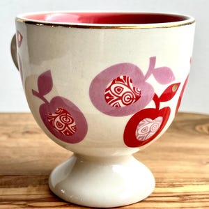 May include: A white ceramic mug with a red rim and gold trim. The mug has a pink and red apple design with a white background. The mug has a pedestal base.