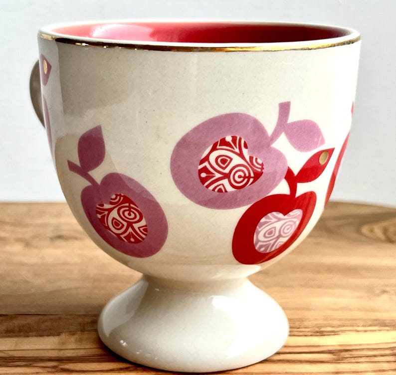 May include: A white ceramic mug with a red rim and gold trim. The mug has a pink and red apple design with a white background. The mug has a pedestal base.