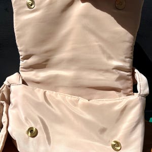 K&ouml;nnte beinhalten: Offene Ansicht einer beige gesteppten Handtasche mit Klappenverschluss. Die Tasche hat ein weiches, gepolstertes Design und goldfarbene Magnetverschl&uuml;sse. Das Innenfutter ist in passendem Beige gehalten.