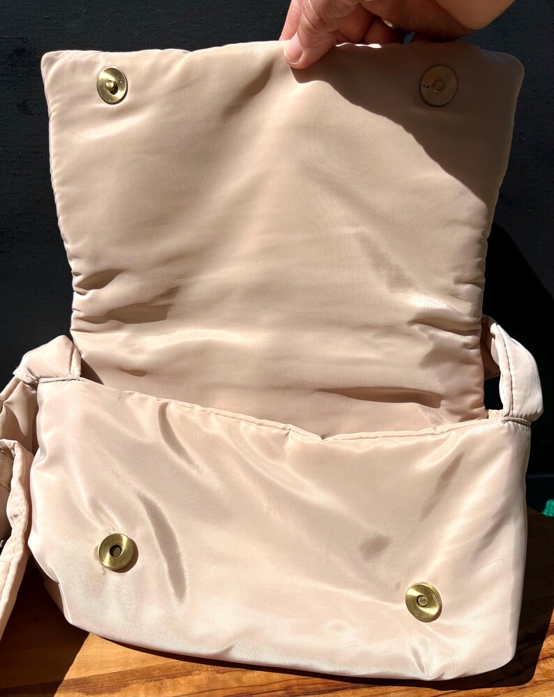 K&ouml;nnte beinhalten: Offene Ansicht einer beige gesteppten Handtasche mit Klappenverschluss. Die Tasche hat ein weiches, gepolstertes Design und goldfarbene Magnetverschl&uuml;sse. Das Innenfutter ist in passendem Beige gehalten.