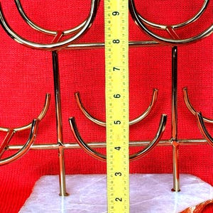 May include: A gold-coloured metal wine glass holder with a white and gold stone base. The holder is approximately 25 cm tall. The holder has a modern design with curved arms to hold the glasses. The background is red.