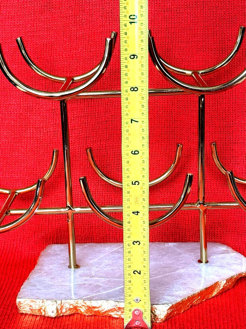 May include: A gold-coloured metal wine glass holder with a white and gold stone base. The holder is approximately 25 cm tall. The holder has a modern design with curved arms to hold the glasses. The background is red.