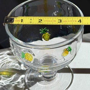 May include: Clear glass footed dessert bowl with yellow and green pineapple designs. The bowl is approximately 4 inches in diameter, as measured by a yellow tape measure.