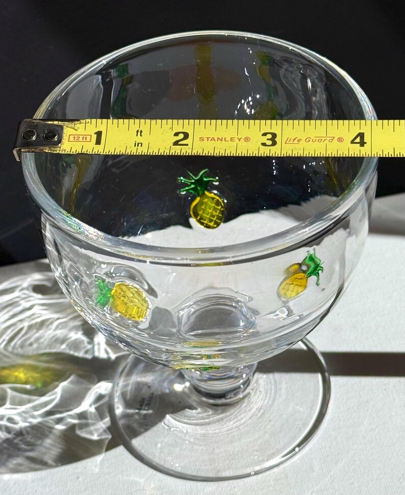 May include: Clear glass footed dessert bowl with yellow and green pineapple designs. The bowl is approximately 4 inches in diameter, as measured by a yellow tape measure.