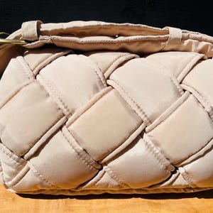 K&ouml;nnte beinhalten: Beige Schultertasche im geflochtenen Stil mit gepolstertem Design. Die Tasche hat einen langen Riemen und eine goldfarbene Schnalle. Die Tasche ist aus weichem, gestepptem Material gefertigt und f&uuml;r den t&auml;glichen Gebrauch geeignet.