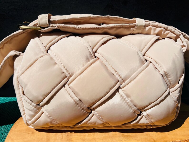 K&ouml;nnte beinhalten: Beige Schultertasche im geflochtenen Stil mit gepolstertem Design. Die Tasche hat einen langen Riemen und eine goldfarbene Schnalle. Die Tasche ist aus weichem, gestepptem Material gefertigt und f&uuml;r den t&auml;glichen Gebrauch geeignet.