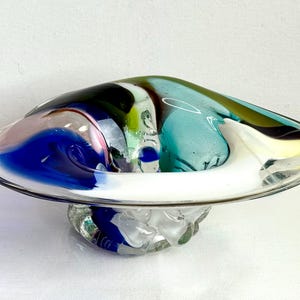 May include: A colorful, hand-blown glass bowl with a wide, shallow design. The bowl features swirls of blue, green, white, and yellow glass. The base is a cluster of clear glass. A decorative piece for the home.