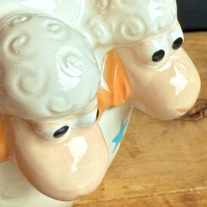 May include: A ceramic sheep-shaped pitcher with a cream-colored body and a light peach face. The sheep has black eyes and orange accents on the ears. A blue star is visible on the side of the pitcher.