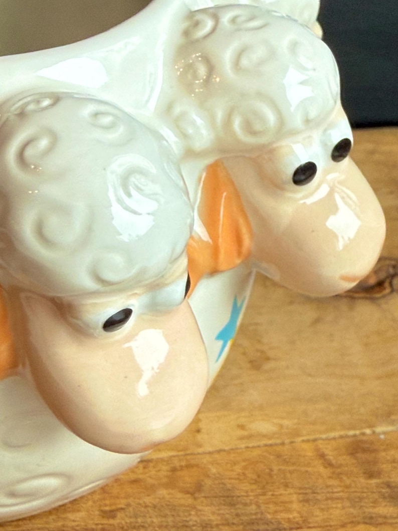 May include: A ceramic sheep-shaped pitcher with a cream-colored body and a light peach face. The sheep has black eyes and orange accents on the ears. A blue star is visible on the side of the pitcher.
