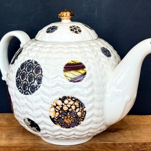 May include: White ceramic teapot with a textured surface and decorative, patterned circles in various colors. The teapot has a curved handle, a spout, and a gold-colored finial on the lid. The teapot is sitting on a wooden surface.