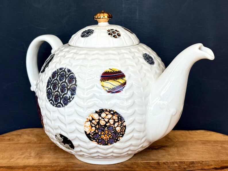 May include: White ceramic teapot with a textured surface and decorative, patterned circles in various colors. The teapot has a curved handle, a spout, and a gold-colored finial on the lid. The teapot is sitting on a wooden surface.