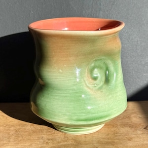 May include: Handmade ceramic vase with a unique shape and color gradient. The interior is a vibrant coral, transitioning to a warm tan and then a soft green. A decorative swirl is on the side.
