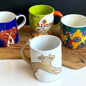 May include: Four decorative ceramic mugs. One is blue with a dog portrait, another is green with a bird design, a third is teal with a cat in a hat, and the last is white with a cat illustration. Each mug has a unique handle design.