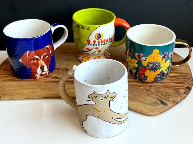 May include: Four decorative ceramic mugs. One is blue with a dog portrait, another is green with a bird design, a third is teal with a cat in a hat, and the last is white with a cat illustration. Each mug has a unique handle design.