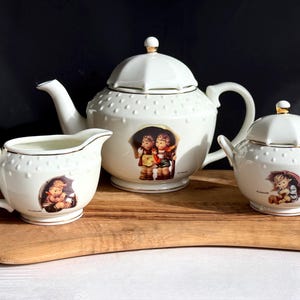 May include: A white porcelain tea set with a teapot, creamer, and sugar bowl. Each piece features a charming illustration of children under an umbrella and is accented with gold trim. The teapot has a lid with a gold knob.