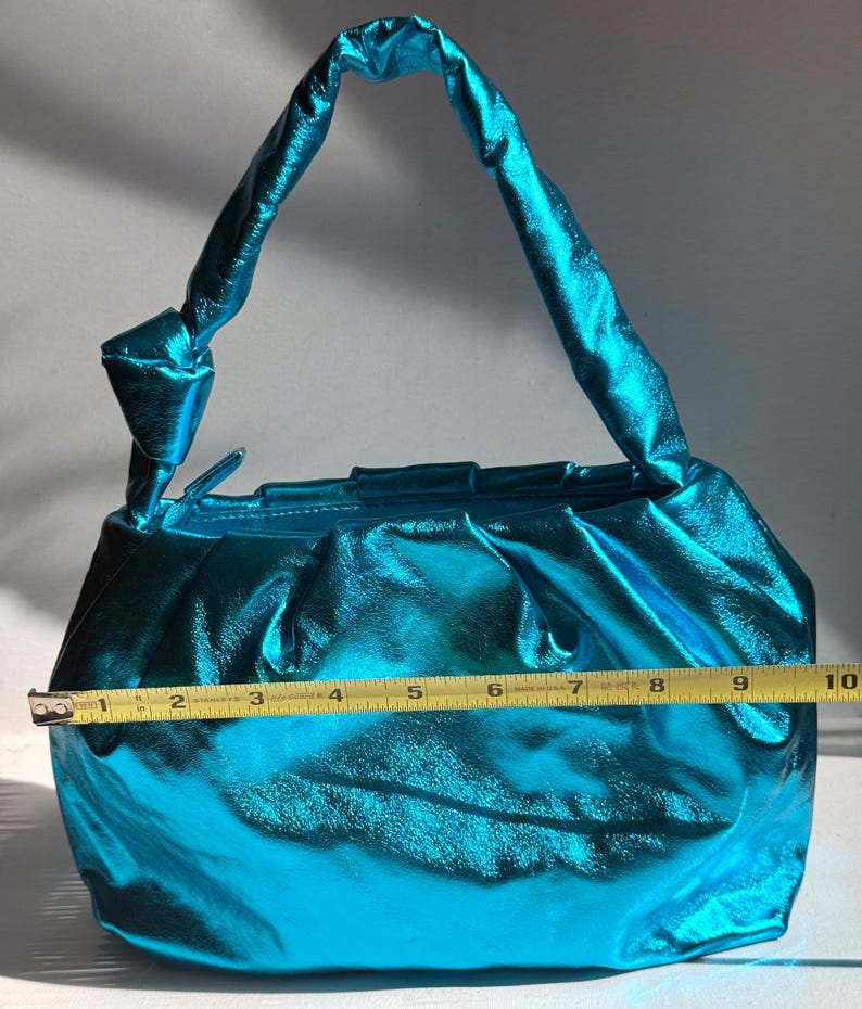 May include: A shiny, turquoise handbag with a knotted handle. The bag is approximately 10 inches wide, with a gathered design and a metallic sheen. The handle is also turquoise and knotted at the base.